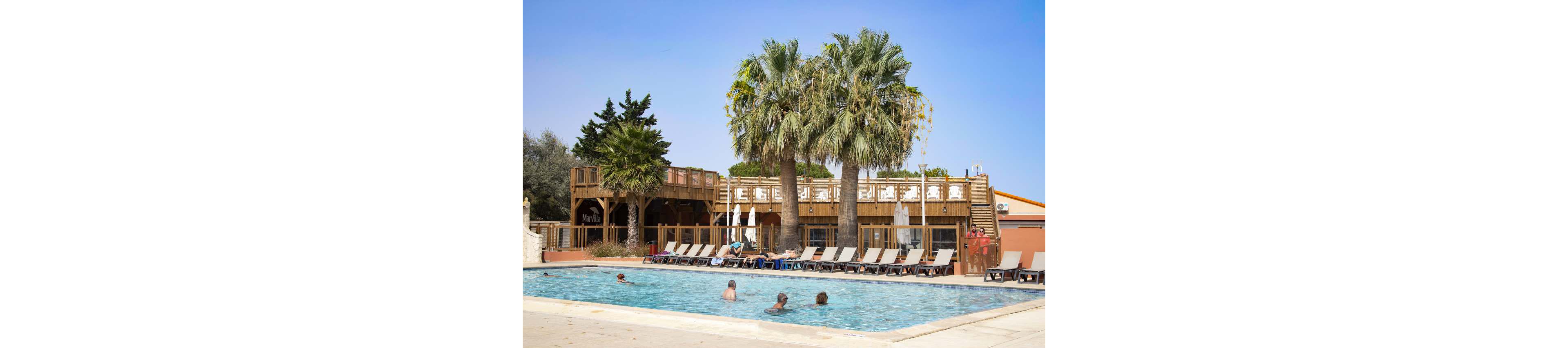 An outdoor swimming pool is surrounded by lounge chairs at a resort. Several people relax in the water while others sunbathe on the deck. Tall palm trees and wooden terraces create a tropical, vacation atmosphere. The sky is clear and blue, suggesting a warm, sunny day.