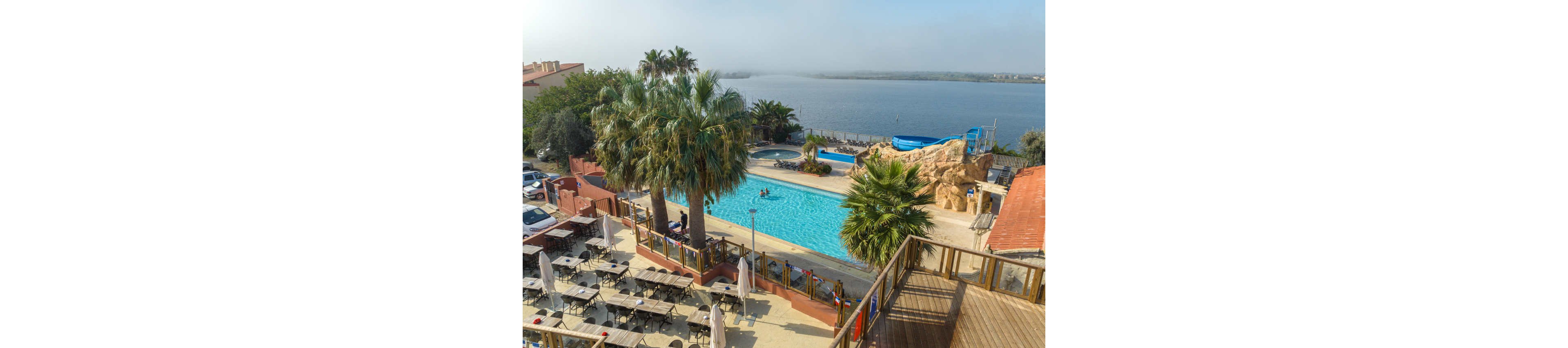 An elevated view shows a resort swimming pool surrounded by palm trees near a calm lake. Sun loungers and closed umbrellas line the pool deck, while an adjacent terrace holds rows of empty tables and chairs. A blue waterslide curves into a smaller pool beside rocky landscaping. Parked cars and low buildings frame the scene under a hazy sky, suggesting a quiet, off-peak moment at the holiday destination.