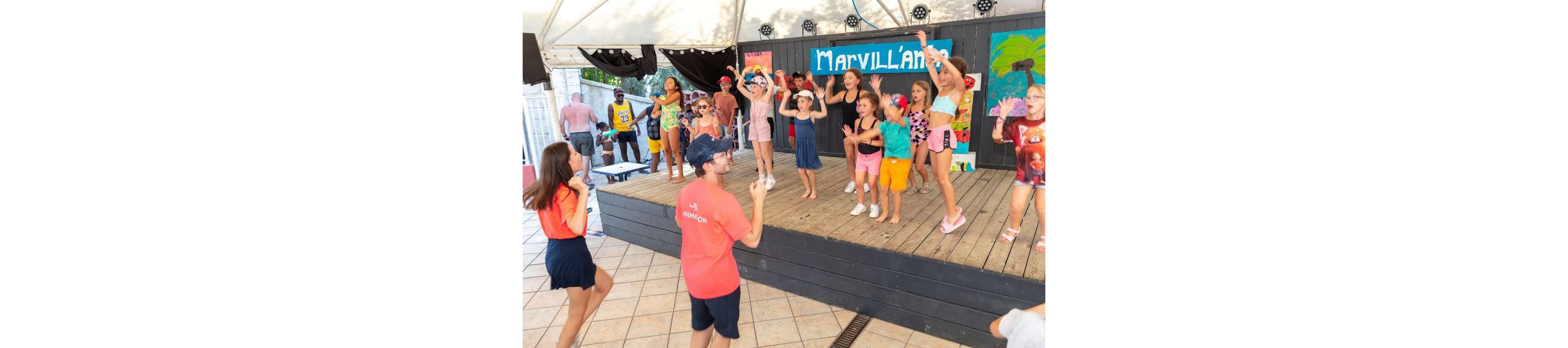 A group of children dance and play on a small wooden stage during an organized activity. Two adult entertainers in red shirts lead the group from the front and side, encouraging participation. The setting appears to be a sunny vacation resort with a covered stage area and colorful decorations. A bright sign reading "Marvilland" and painted backdrops add a festive, playful atmosphere.