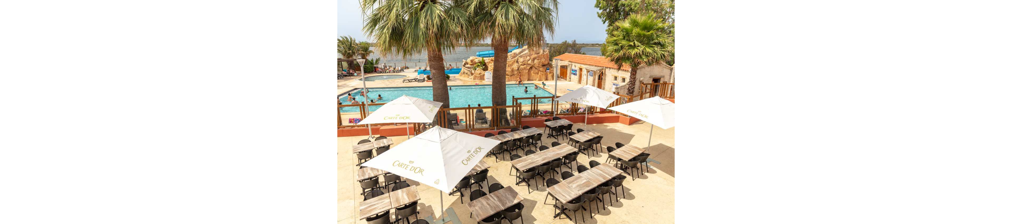 An outdoor café terrace overlooks a swimming pool area on a sunny day. Rows of empty tables and black chairs sit beneath large white umbrellas printed with the words "CAFÉ D'OR". Beyond the terrace, people relax and play in the pool near rock formations and a waterslide. Palm trees and a distant waterfront create a relaxed vacation atmosphere.