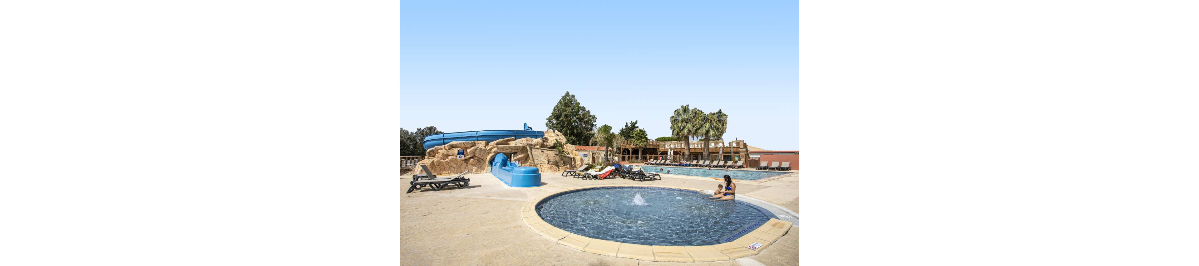 An outdoor resort pool complex features a small circular wading pool in the foreground where an adult and child sit at the water’s edge. Behind them, a larger swimming pool is lined with sun loungers and shaded seating areas. A blue water slide built into artificial rock formations adds a playful attraction to the scene. Palm trees and clear blue sky create a relaxed, sunny vacation atmosphere.
