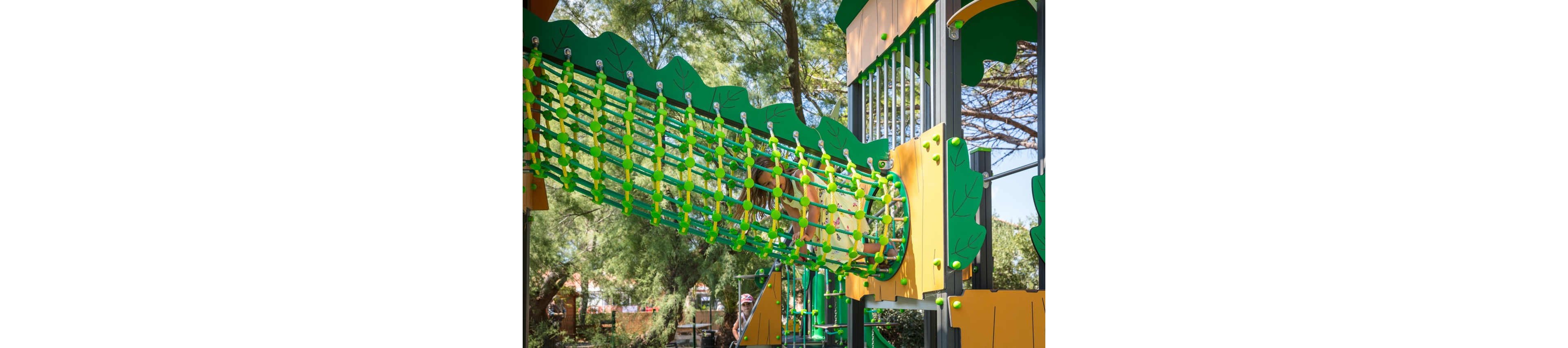 A vibrant green and yellow rope tunnel bridge is featured in an outdoor playground. The structure is surrounded by trees, creating a shaded and natural setting. A child wearing a helmet is partially visible in the background, suggesting active play. The design includes leaf-shaped panels and bright connectors that emphasize a fun, nature-inspired theme.