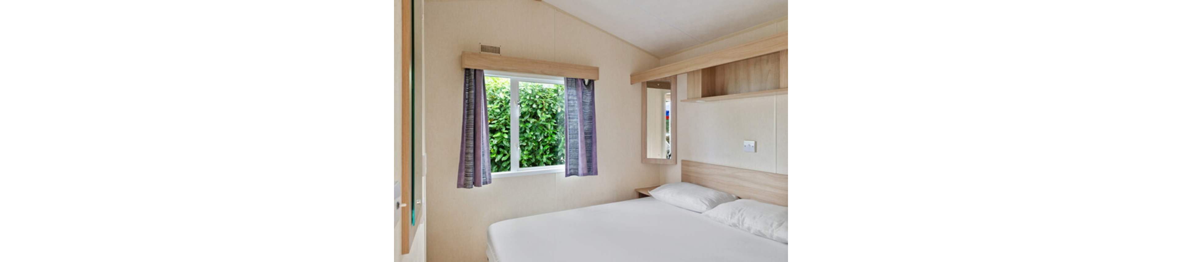 A small, minimalist bedroom features a neatly made double bed with white bedding. A large window with purple curtains lets in natural light and offers a view of dense green foliage outside. Light wood built-in shelves and a mirror add storage and functionality to the compact space. The neutral walls and simple decor create a clean, calm, and uncluttered atmosphere.