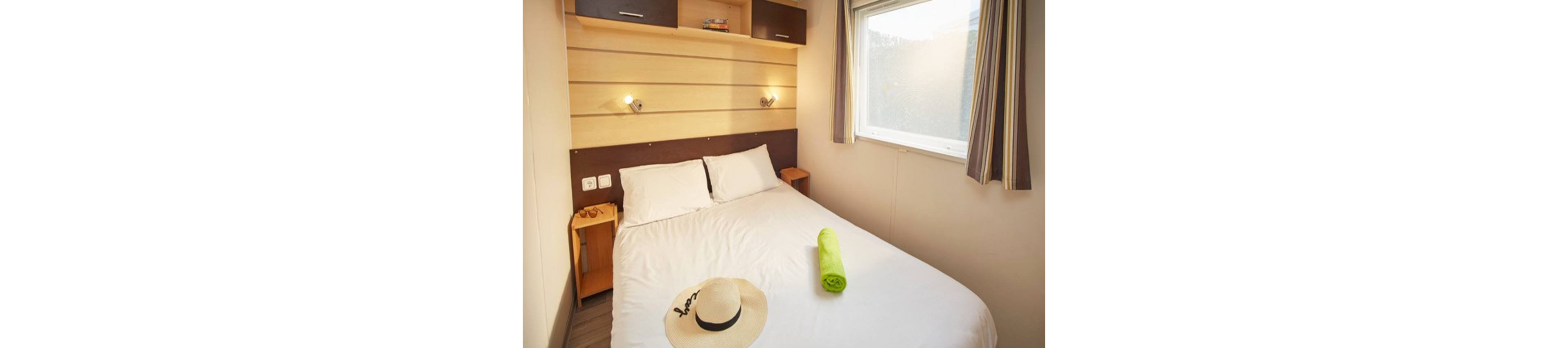 A small, cozy bedroom features a neatly made double bed with white linens and two pillows. A rolled green towel and a straw hat with sunglasses rest on the bed, suggesting a vacation or getaway. Wooden wall panels, bedside tables, and overhead storage cabinets add warmth to the space. Natural light streams in through a large window with striped curtains, creating a bright and inviting atmosphere.