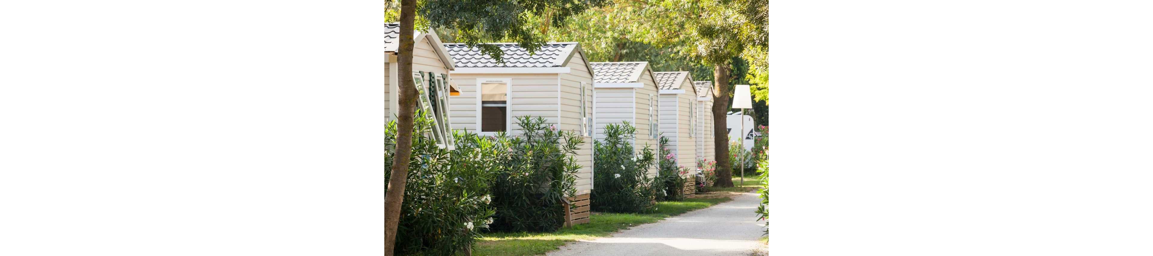 A row of small mobile homes is lined up along a narrow path in a leafy outdoor setting. The light-colored cabins feature windows, pitched roofs, and simple siding. Lush green shrubs and trees surround the homes, creating a peaceful, shaded atmosphere. Bright sunlight filters through the foliage, suggesting a warm, relaxing vacation environment.