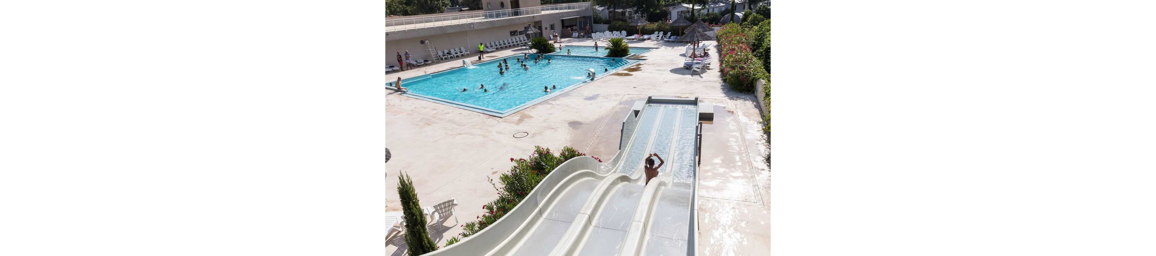 An outdoor leisure complex features a large rectangular swimming pool surrounded by lounge chairs and sunbathers. Several people are swimming and playing in the pool while others relax on the deck. In the foreground, a person rides down a curved waterslide next to a straight slide that ends in a shallow run-out. The bright daylight, clear water, and greenery around the deck create a sunny, vacation-like atmosphere.