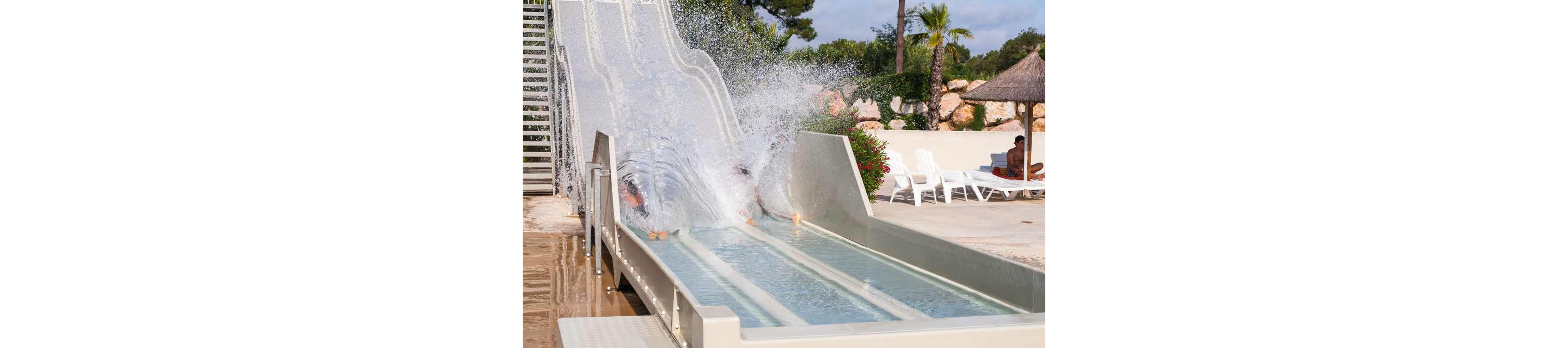 Two people are captured mid-splash as they exit a twin-lane waterslide into a shallow pool. The scene takes place in an outdoor water park with lounge chairs and umbrellas nearby. Water sprays dramatically upward, partially obscuring the riders. Palm trees and rocks in the background suggest a warm, resort-like environment.