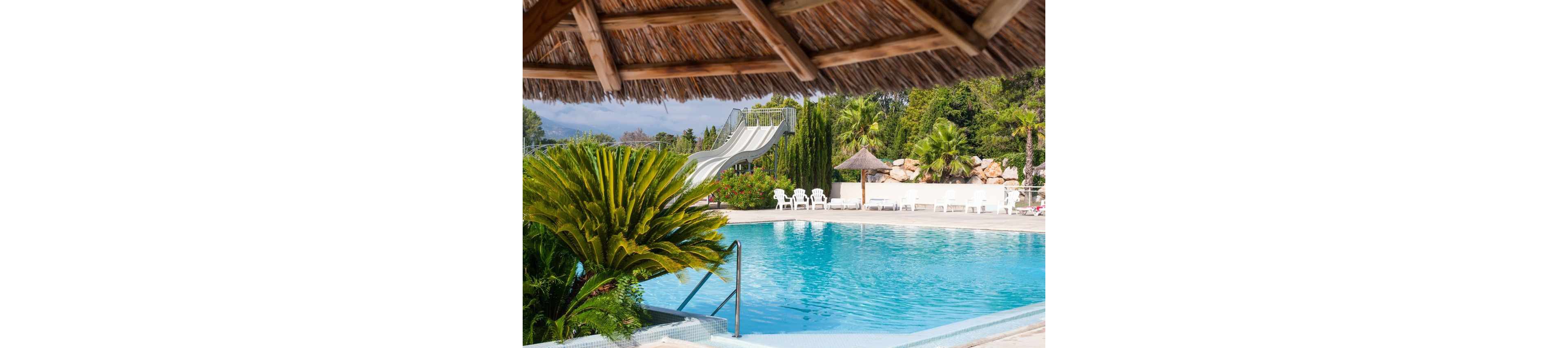 A clear blue swimming pool is framed by lush tropical plants and a thatched roof shade structure. In the background, a tall white waterslide curves down into the pool area. White lounge chairs and umbrellas line the pool deck, surrounded by palm trees and greenery. The bright daylight and calm water create a relaxing vacation atmosphere.