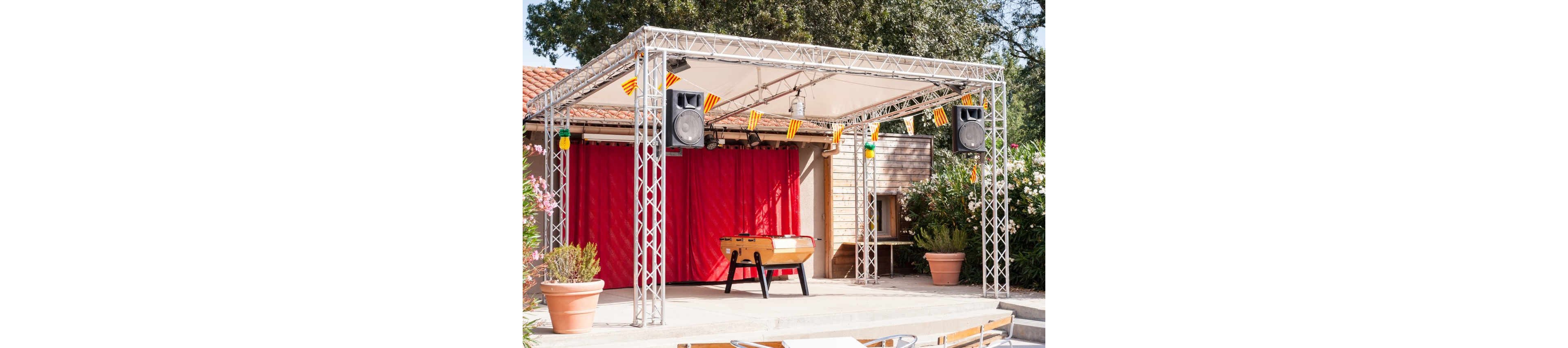 An outdoor stage setup features a foosball table placed at the center beneath a metal truss canopy. Large speakers hang from the structure, and small striped pennant flags decorate the front edge. Behind the stage, bright red curtains cover the backdrop, with potted plants and flowering bushes framing the scene. The setting appears to be a casual entertainment or event area in a sunny courtyard.