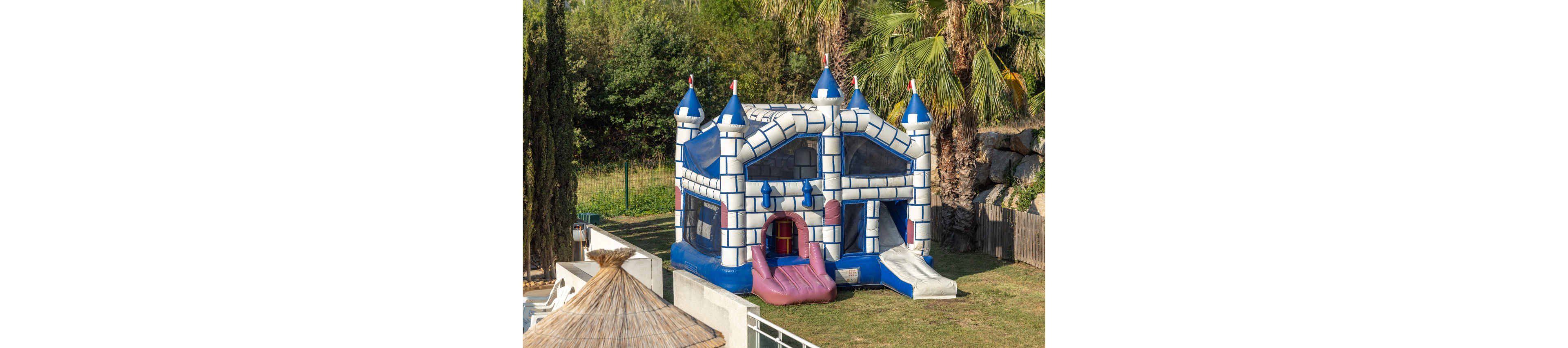 A large inflatable castle bounce house stands on a grassy lawn. The structure is designed like a blue and white stone fortress with turrets and mesh windows. A pink entrance ramp and a white slide extend from the front of the bouncer. Palm trees, shrubs, and a fence surround the play area, suggesting an outdoor recreational or party setting.