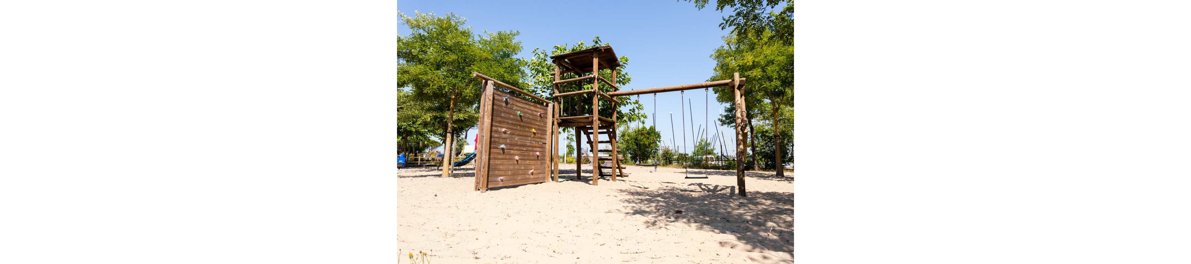 A wooden playground structure with a climbing wall and tower stands on a sandy surface. Several swings hang from a long wooden beam extending from the tower. Green trees surround the play area under a clear blue sky. The playground appears empty and sunlit, suggesting a warm, peaceful day outdoors.