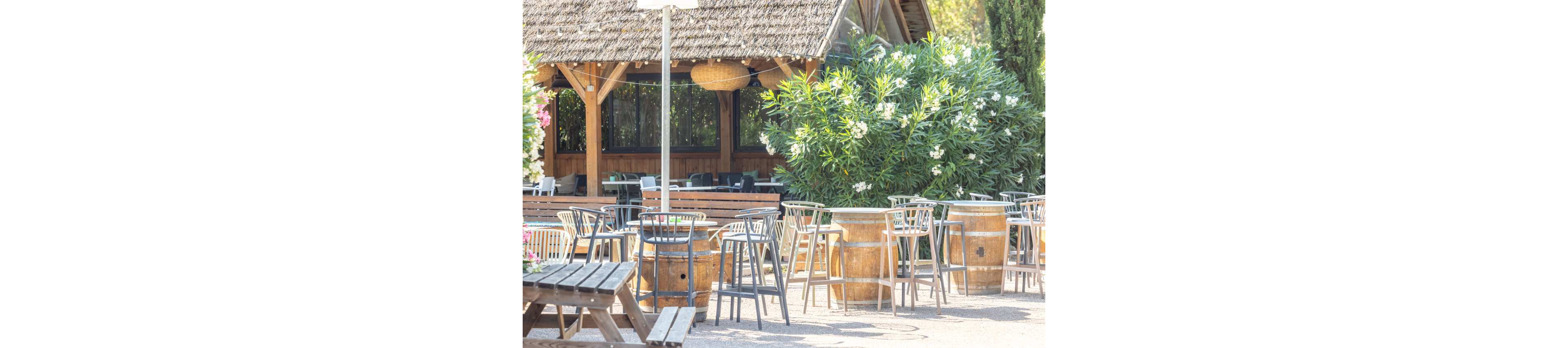 An inviting outdoor patio features wooden barrel tables paired with modern metal chairs. The seating area is set in front of a rustic wooden building with a thatched-style roof. Lush green shrubs with white flowers add natural color and texture to the scene. Bright sunlight and soft shadows create a warm, relaxed atmosphere suitable for dining or social gatherings.