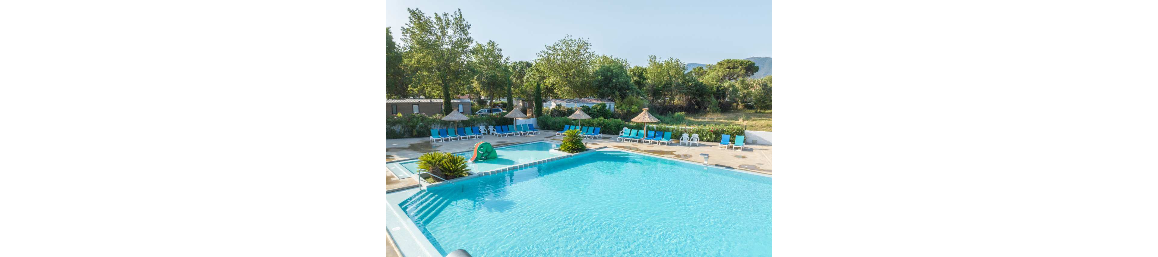 Large clear blue swimming pools sit in an outdoor leisure area surrounded by trees and greenery. Numerous sun loungers line the poolside, some shaded by thatched parasols. A small green slide shaped like an animal adds a playful touch to the shallow pool. The scene is bright and calm, suggesting a relaxing holiday atmosphere in warm weather.
