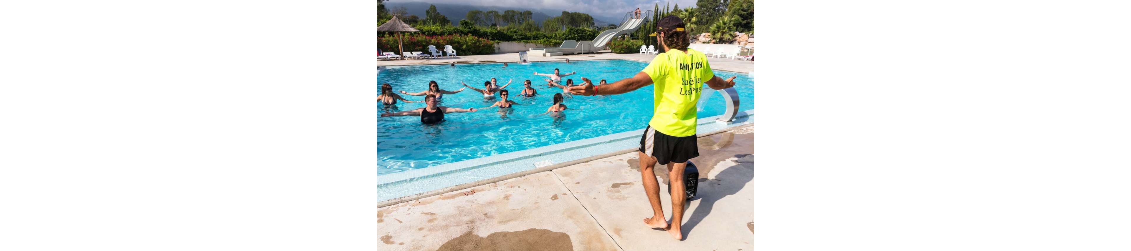 A group of adults participates in a water aerobics class in an outdoor swimming pool. The instructor stands on the pool deck facing the group, demonstrating arm movements. Participants are spaced across the pool, following along in shallow water. Lounge chairs, greenery, and a waterslide are visible in the background, suggesting a resort or holiday setting. The atmosphere is active and social under bright daylight.