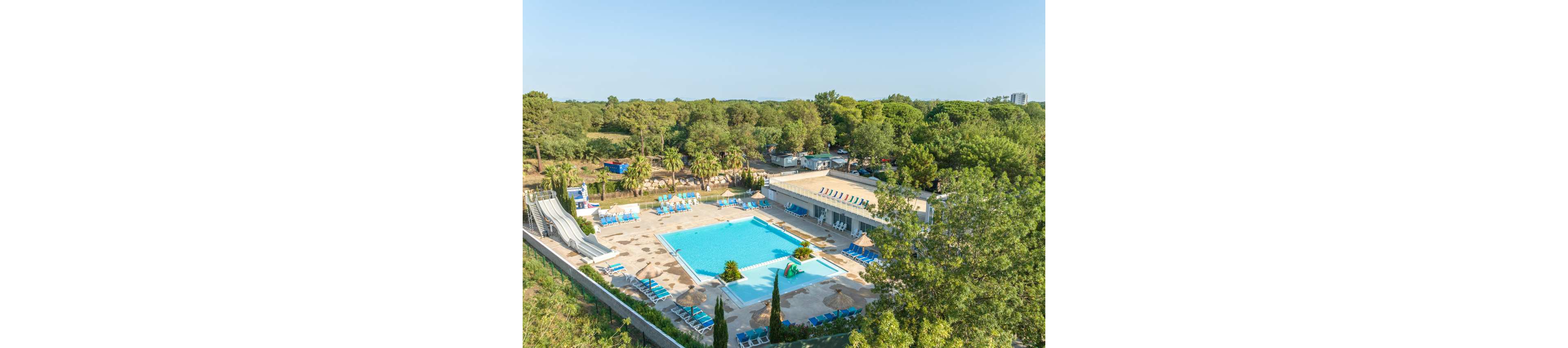 An outdoor swimming pool complex is seen from above, surrounded by sun loungers and parasols. The pool area includes a main rectangular pool and a smaller shallow section, with water slides on one side. The facility is set within a lush green landscape of trees and vegetation, suggesting a holiday resort or campground. Clear blue skies and bright sunlight create a relaxed, summery atmosphere.