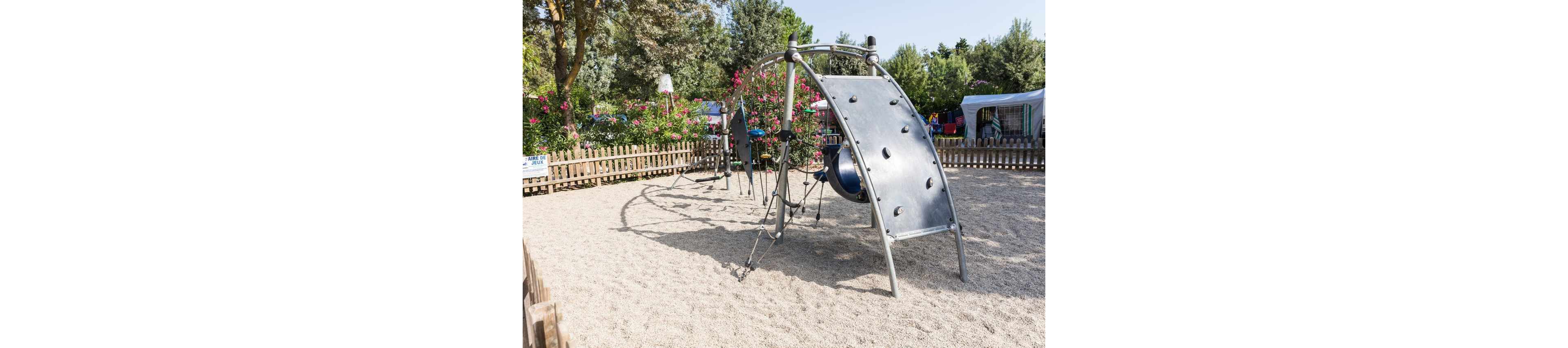 A contemporary metal climbing structure stands in the middle of a sandy playground. The equipment features curved frames, ropes, and a climbing wall panel. The play area is enclosed by a wooden fence with flowering bushes and trees in the background. Tents or caravans are visible beyond the fence, suggesting a campsite or recreational park setting under clear daylight.