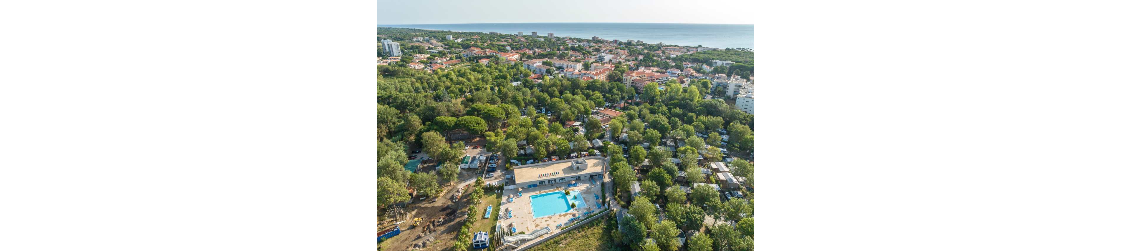 A large outdoor swimming pool complex is surrounded by trees in a lush green resort. The aerial view reveals numerous buildings, campsites, and facilities nestled within the forested area. In the distance, a coastal town stretches toward the shoreline and the calm sea. The scene suggests a popular holiday destination combining nature, recreation, and seaside access.