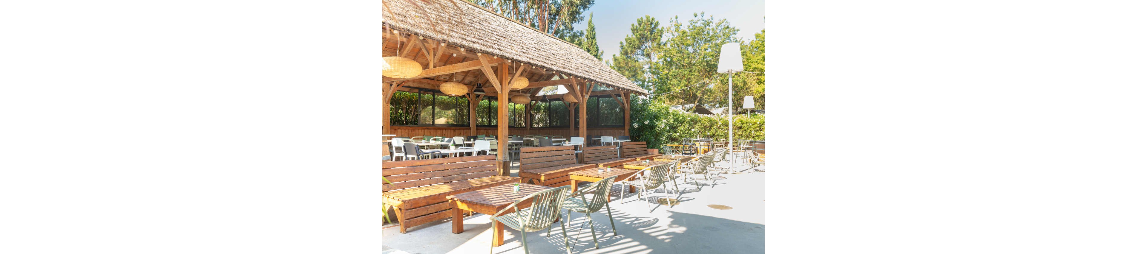 An outdoor patio area features long wooden benches and tables arranged in rows under a rustic wooden pergola. The space is bright and sunlit, with modern metal chairs and additional seating extending into the open courtyard. Large woven pendant lights hang from the pergola ceiling, adding a warm decorative touch. Surrounding greenery and trees create a relaxed, natural atmosphere suitable for dining or social gatherings.
