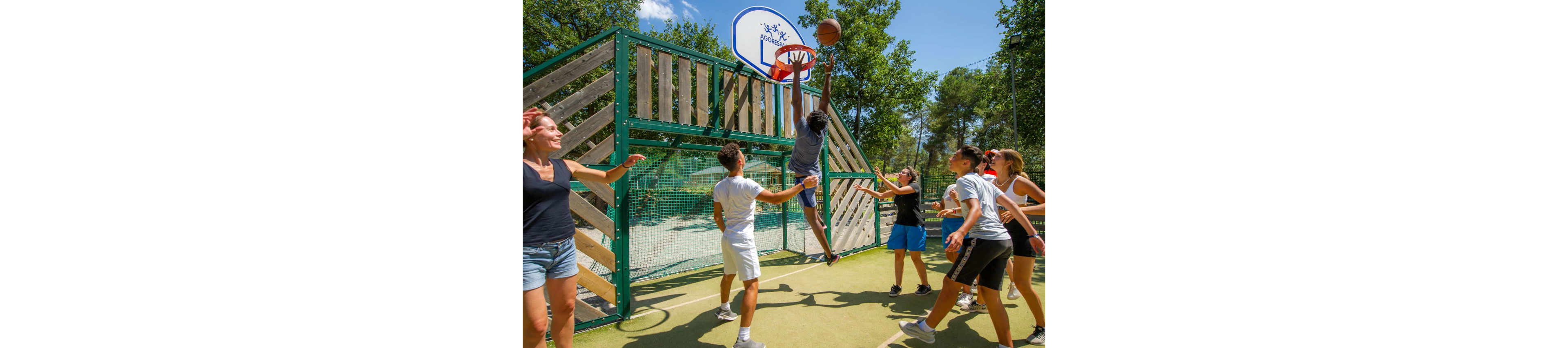 A group of teenagers play an energetic game of basketball on an outdoor court. One player jumps high to make a shot while others reach up to defend or watch. The court is surrounded by a wooden and metal fence with a hoop mounted above. Trees and a bright blue sky create a lively summer atmosphere. The scene conveys teamwork, recreation, and active youth sports.