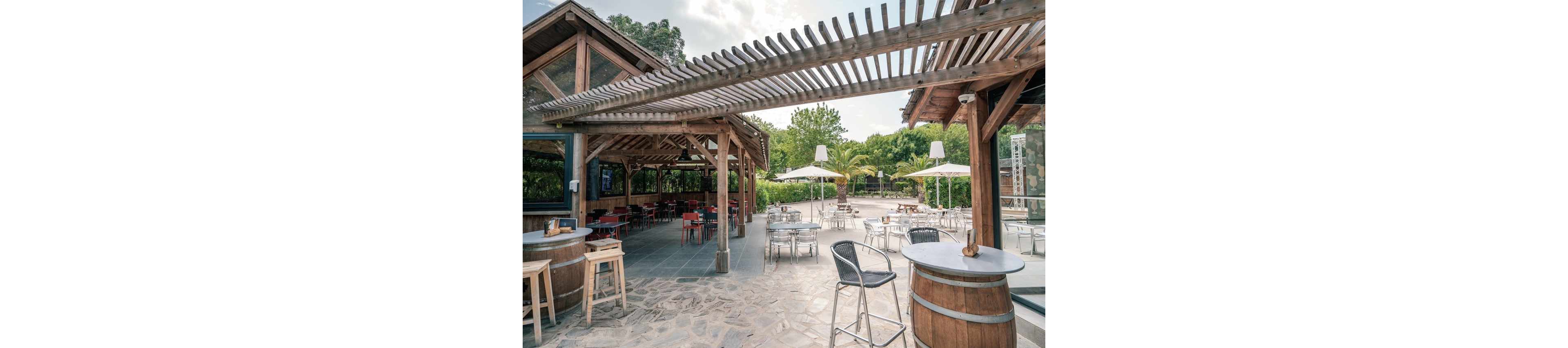An outdoor bar area features wooden pergolas and rustic barrel tables on a stone-paved terrace. Metal chairs and round tables are arranged across the open space, with additional seating under large white umbrellas. The surrounding greenery and palm trees create a relaxed, resort-like atmosphere. The scene appears quiet and empty, suggesting early opening hours or an off-peak time.
