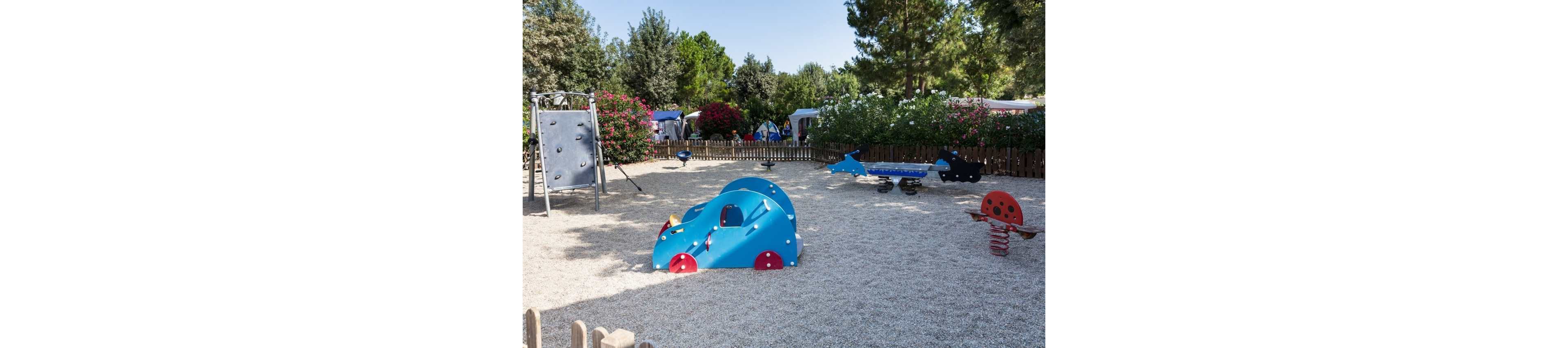 An empty outdoor playground features several colorful play structures on a gravel surface. In the foreground, a blue car-shaped climber and a red ladybug spring rider stand out. Additional equipment, including a small climbing wall and animal-shaped seesaws, are visible in the background. The area is enclosed by a wooden fence and surrounded by trees, bushes, and nearby tents or mobile homes under a clear sky.