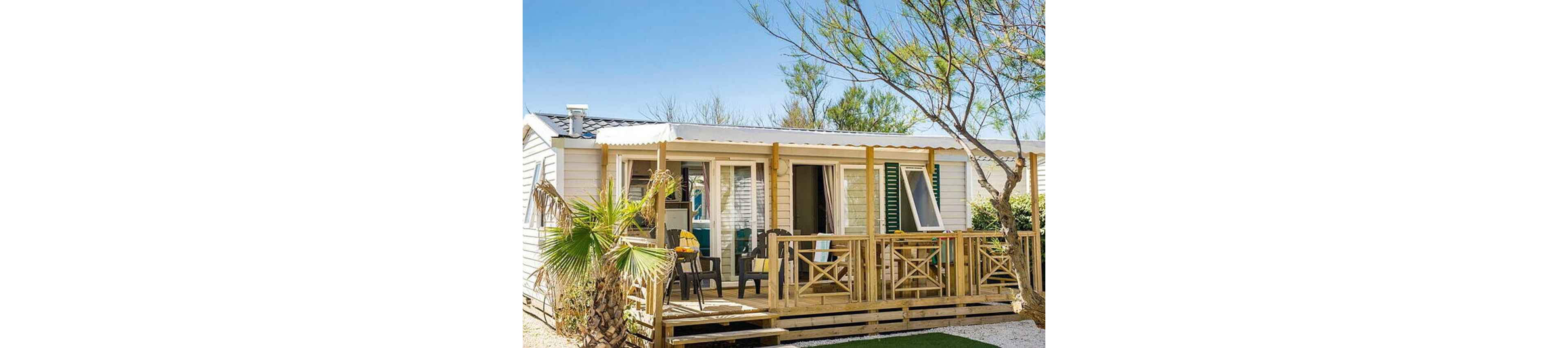 A bright mobile home with a spacious wooden terrace is shown in a sunny outdoor setting. Several chairs and a table are arranged on the deck, suggesting a relaxed vacation atmosphere. The structure features white siding, green shutters, and large windows that open onto the patio. Palm trees and other greenery surround the home, enhancing the warm, coastal holiday feel.