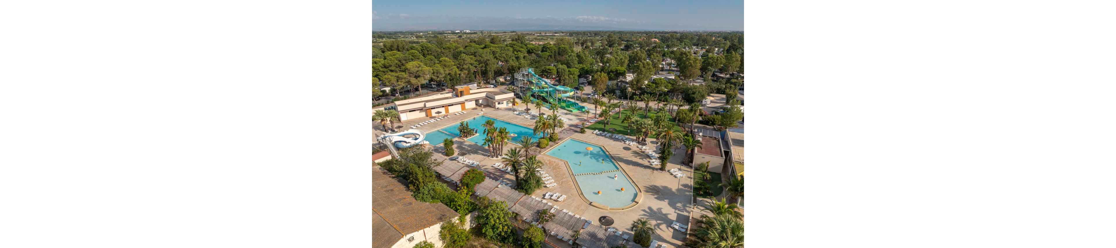A large outdoor resort complex features multiple swimming pools and winding water slides surrounded by palm trees. The scene is captured from above, showing sun loungers neatly arranged around the pools and open deck areas. A shallow, irregularly shaped pool with small play features sits beside a deeper rectangular pool. Beyond the water park, a dense line of trees and low buildings stretches into the distance under a clear sky.
