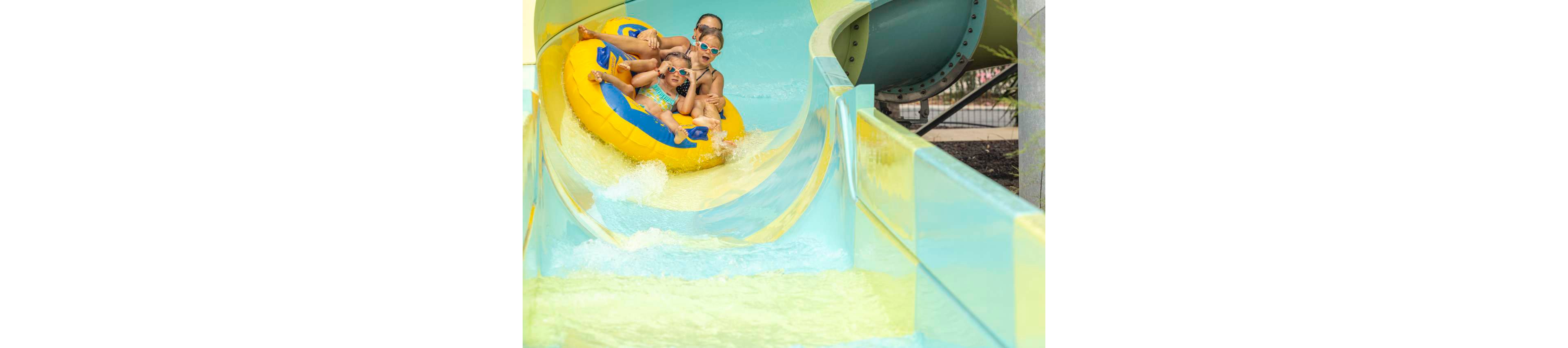 A group of people ride together on a large yellow inflatable raft down a curving water slide. The slide features bright blue and green sections with splashing water around the raft. The riders appear to be enjoying a fun, fast descent at an outdoor water park. Structural elements of the slide and surrounding park are visible in the background.