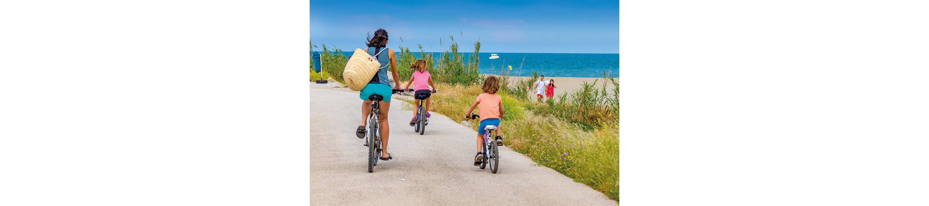 A woman and two children ride bicycles along a paved path near the sea. Tall grasses line the trail, and a sandy beach with people walking is visible in the background. The sky is bright blue, and a small boat can be seen on the water. The scene suggests a relaxed summer vacation and outdoor family activity by the coast.
