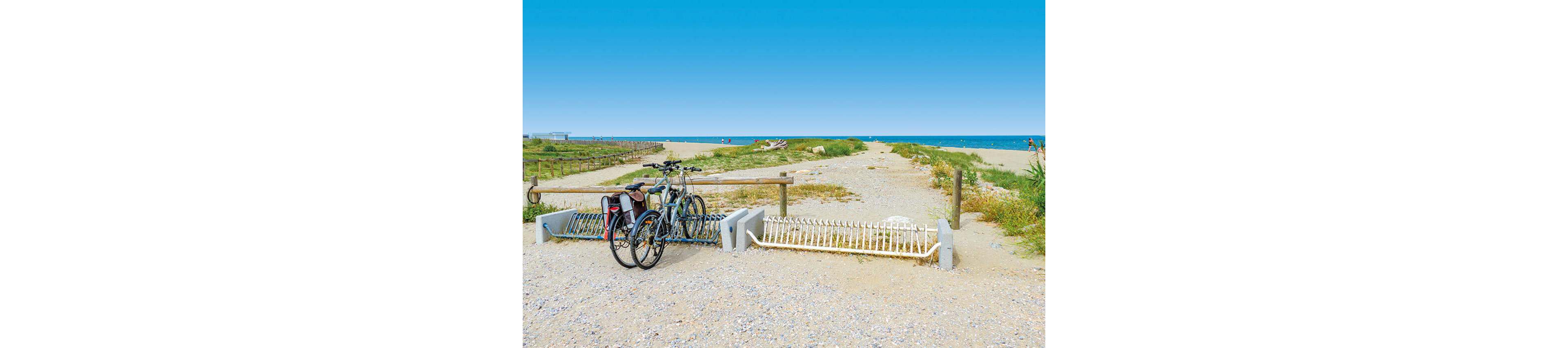 Two bicycles are parked at the entrance of a sandy path leading to the beach. The scene opens toward the sea under a clear blue sky, with dunes and low vegetation on either side of the trail. A simple wooden fence and white metal barriers frame the pathway. The distant shoreline and calm water create a relaxed, summery coastal atmosphere.