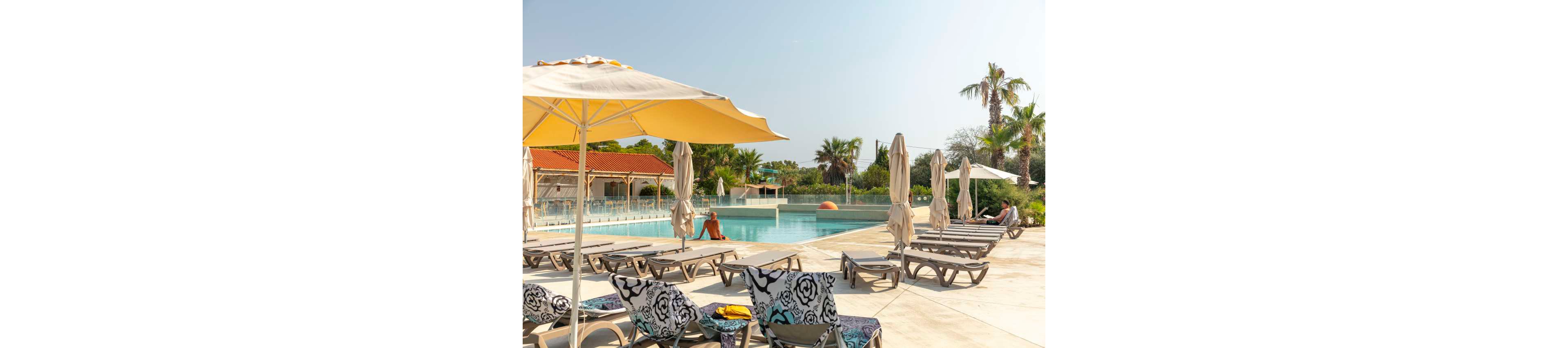 An outdoor resort swimming pool is surrounded by empty loungers and large sun umbrellas on a bright day. A few people relax at the poolside, enjoying the calm atmosphere and clear blue water. Palm trees and greenery frame the scene, with low buildings and a bar area in the background. The overall mood is tranquil and summery, suggesting a warm vacation destination.