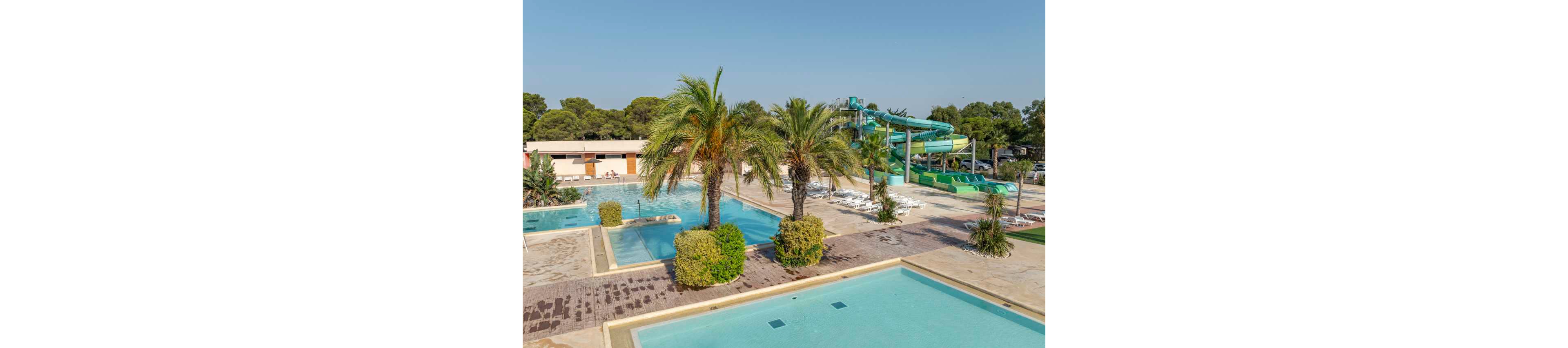 An outdoor water park features several clear blue swimming pools surrounded by palm trees and low shrubs. In the background, large green water slides curve above the pool deck, with rows of white lounge chairs nearby. The scene is set on a sunny day with a clear blue sky and dense trees beyond the park. The overall atmosphere is relaxed and inviting, suggesting a family-friendly vacation destination.