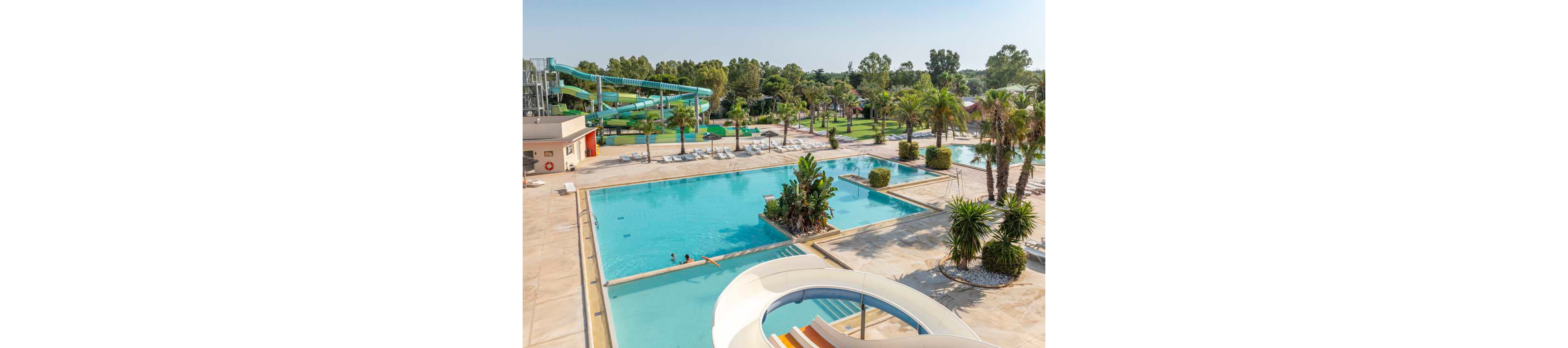 Large outdoor swimming pools are shown under a clear sunny sky at a resort-style water park. Multiple pools of varying depths are surrounded by lounge chairs, palm trees, and landscaped greenery. A curved white waterslide and a tall green tube slide structure dominate the background, adding a sense of fun and activity. A few people relax in the water, emphasizing a family-friendly vacation atmosphere.