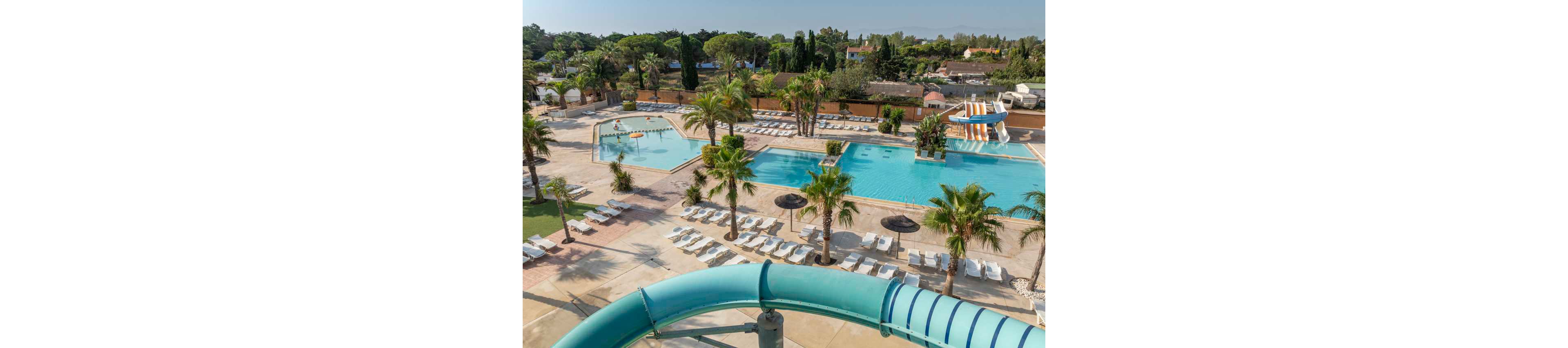 A large outdoor resort pool complex is shown from above on a sunny day. Multiple swimming pools of varying depths are surrounded by rows of white lounge chairs and palm trees. A curved turquoise waterslide enters the scene in the foreground, with another slide structure visible near the main pool. The area appears calm with only a few people in the water, creating a relaxed vacation atmosphere.