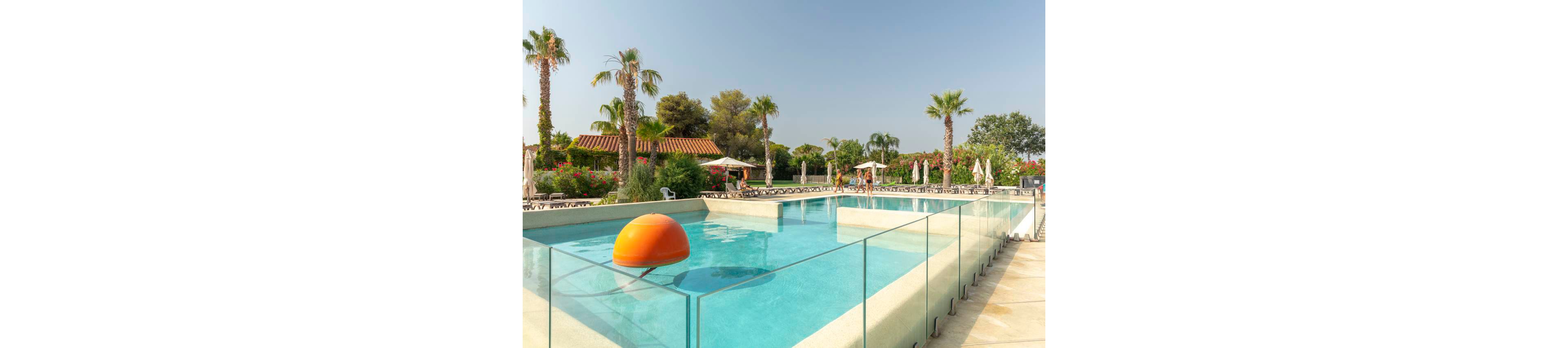 An outdoor swimming pool is surrounded by clear glass barriers at a sunny resort. A large orange floating ball sits in the water, adding a playful accent. Palm trees, sun loungers, and white parasols line the background near a low building with a tiled roof. The bright daylight and clear sky create a relaxed vacation atmosphere.
