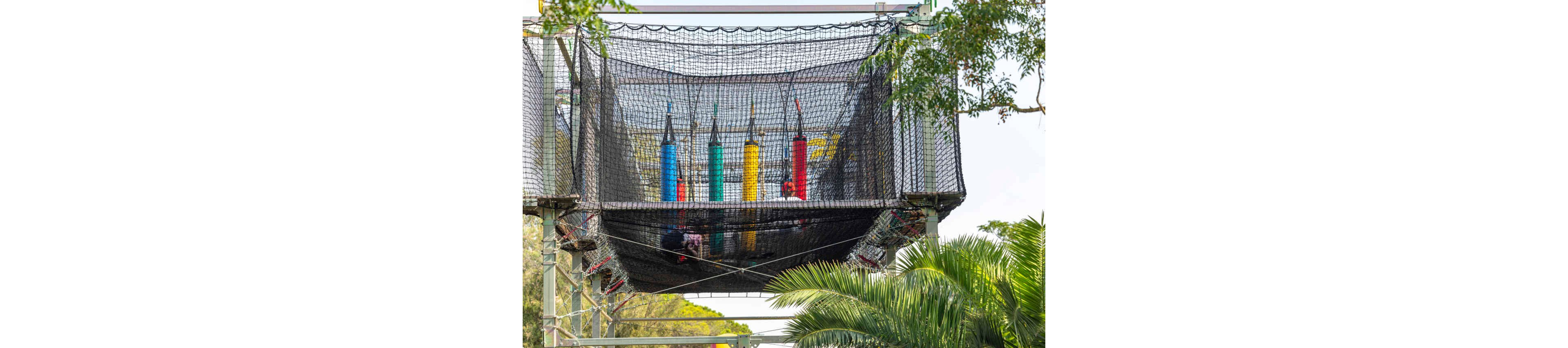 A high ropes obstacle course is suspended in the air, featuring several brightly colored vertical cylinders enclosed within a safety net. The structure is elevated on metal supports and surrounded by lush green trees and palm fronds. The scene suggests an outdoor adventure park or zipline attraction designed for recreational climbing and balancing challenges. The overall atmosphere is playful and energetic with strong primary colors standing out against the natural background.
