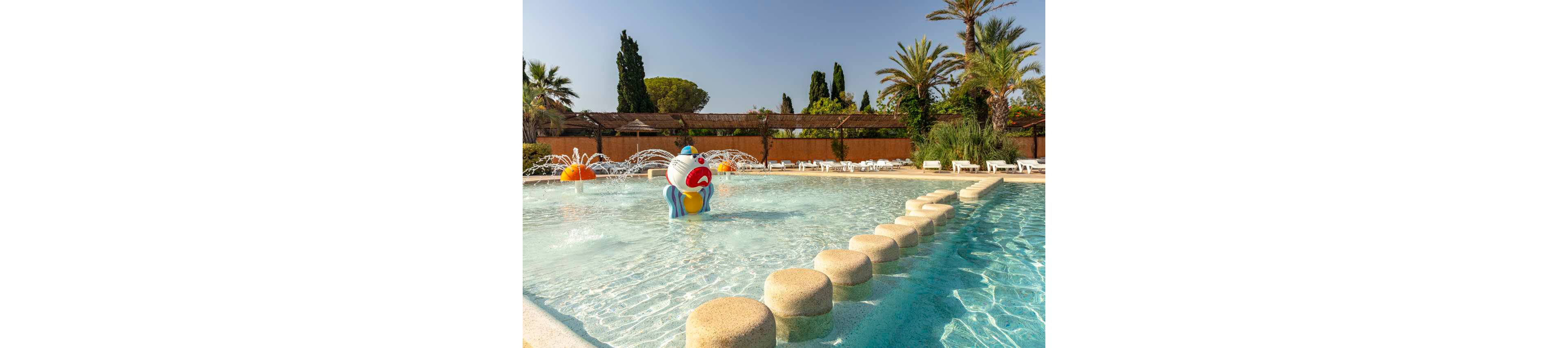 A shallow resort swimming pool features a colorful cartoon character fountain at its center. Round stepping stones lead across the clear water toward the playful structure. In the background, lounge chairs line the poolside beneath wooden pergolas and lush greenery. Tall palm trees and bright sunlight create a warm, inviting vacation atmosphere.