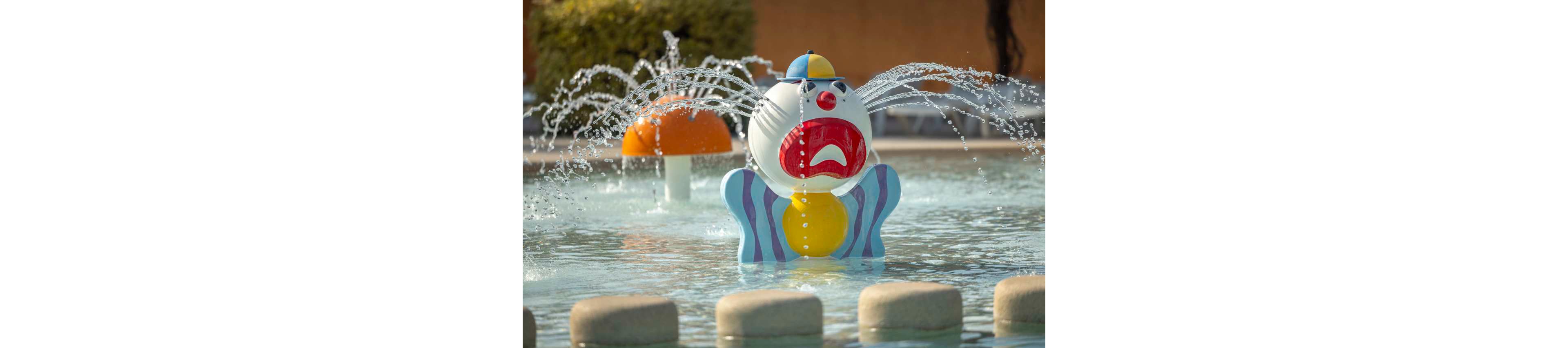 A colorful clown-themed water feature sprays streams of water in a shallow splash pool. The clown figure has a red mouth, blue striped body, and yellow details, creating a playful centerpiece. In the background, a mushroom-shaped fountain and poolside seating are softly out of focus. The scene suggests a family-friendly water park or children's pool area on a sunny day.