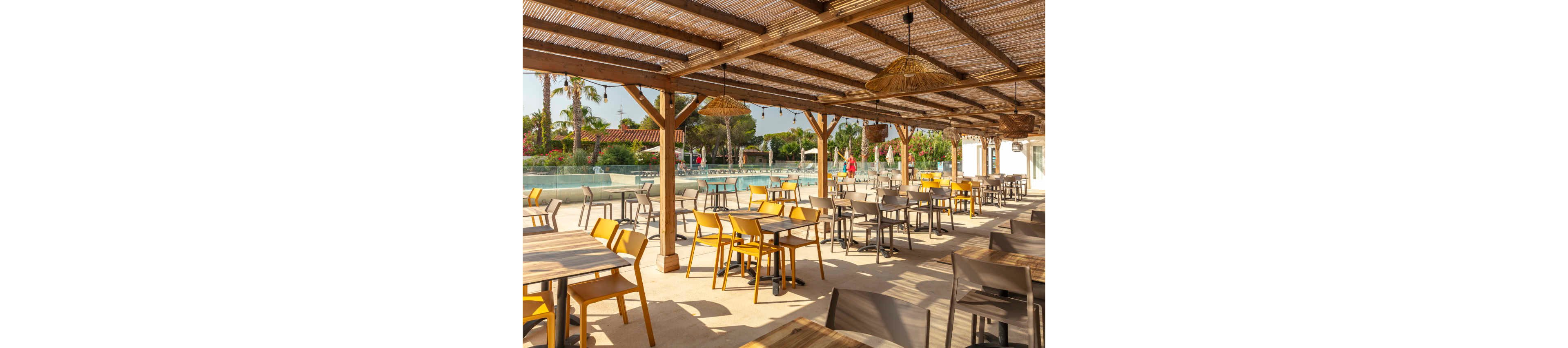 An open-air café terrace with wooden pergola roofing overlooks a large swimming pool. Numerous empty tables and chairs, some yellow and some gray, are neatly arranged in rows. Palm trees, greenery, and low buildings surround the pool area in the background. The bright sunlight and clear sky create a warm, relaxed vacation atmosphere.