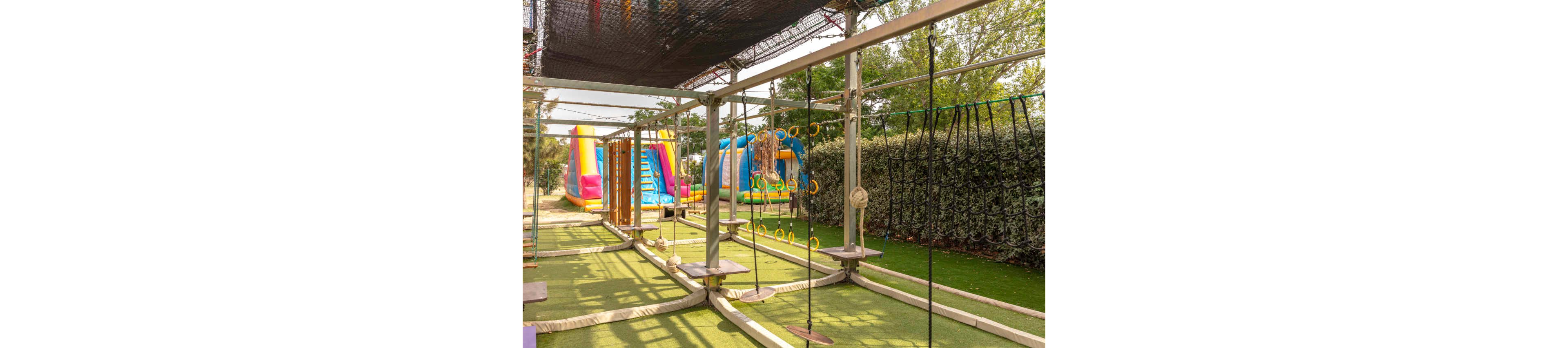 An outdoor adventure playground features a rope obstacle course with hanging platforms, rings, and nets. The structures are set on artificial grass under a metal frame, surrounded by tall hedges and trees. In the background, colorful inflatable slides and bounce houses add a fun, carnival-like atmosphere. The scene is brightly lit, suggesting a sunny day at a family-friendly park.