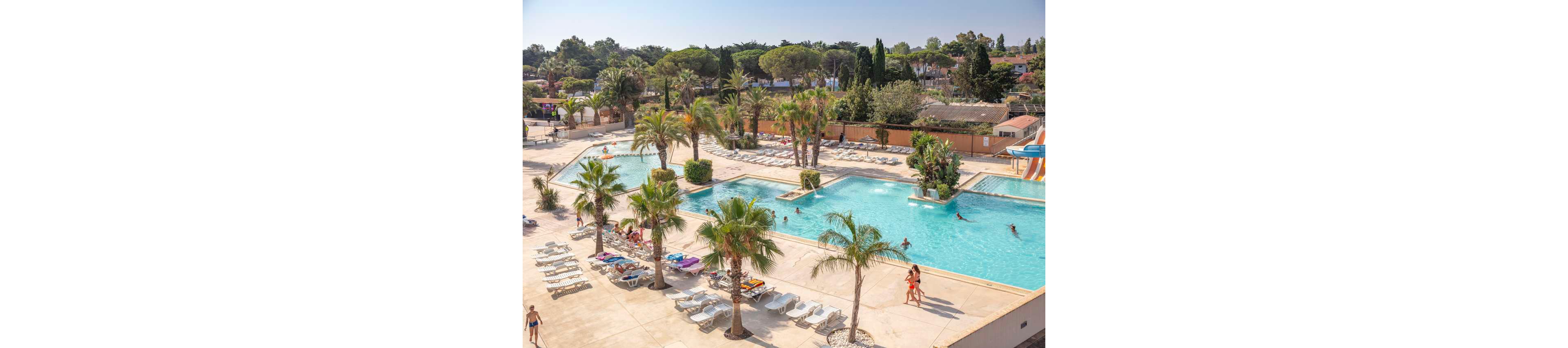 Large outdoor swimming pools are surrounded by sun loungers and tall palm trees at a holiday resort. People relax on chairs and swim in the clear blue water under bright sunlight. A waterslide and additional pool areas are visible in the background, along with low buildings and dense greenery. The scene conveys a warm, leisurely vacation atmosphere.
