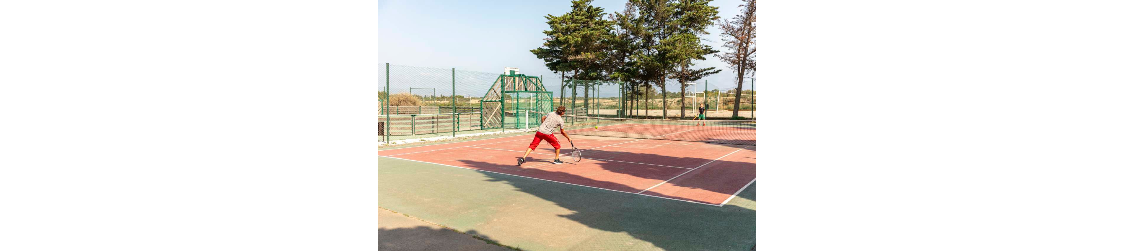 An adult tennis player in red shorts prepares to hit a shot on an outdoor court. Another player stands on the opposite side of the net, ready to return the ball. The scene is set on a sunny day with clear skies, surrounded by tall trees and fencing. Shadows on the court emphasize the bright natural light and open, recreational atmosphere.