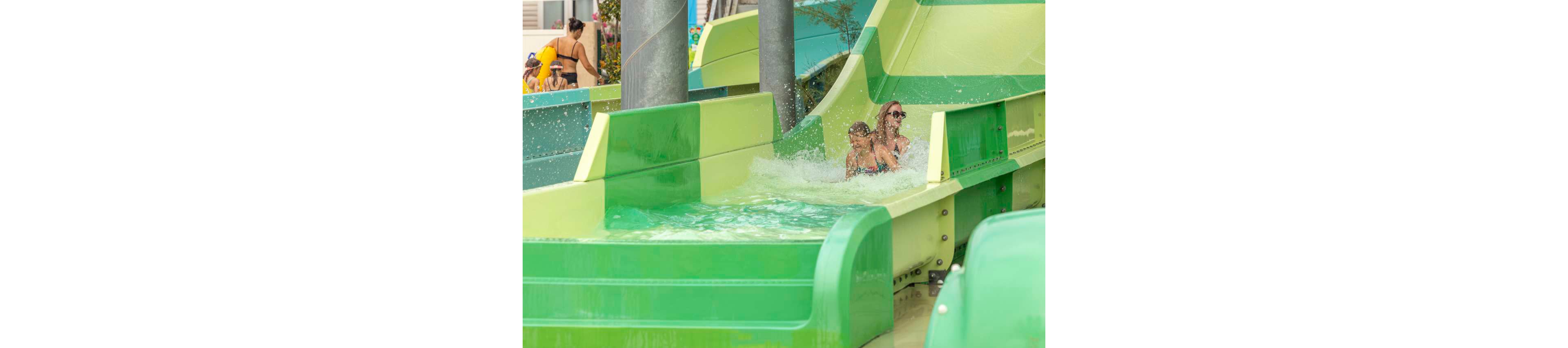 Two children ride together down a bright green waterslide, splashing into the shallow pool at the bottom. The scene takes place at an outdoor water park with other visitors visible in the background. Strong green tones from the slide contrast with the light blue water, creating a playful summer atmosphere. The image captures motion, fun, and family recreation in a warm-weather setting.