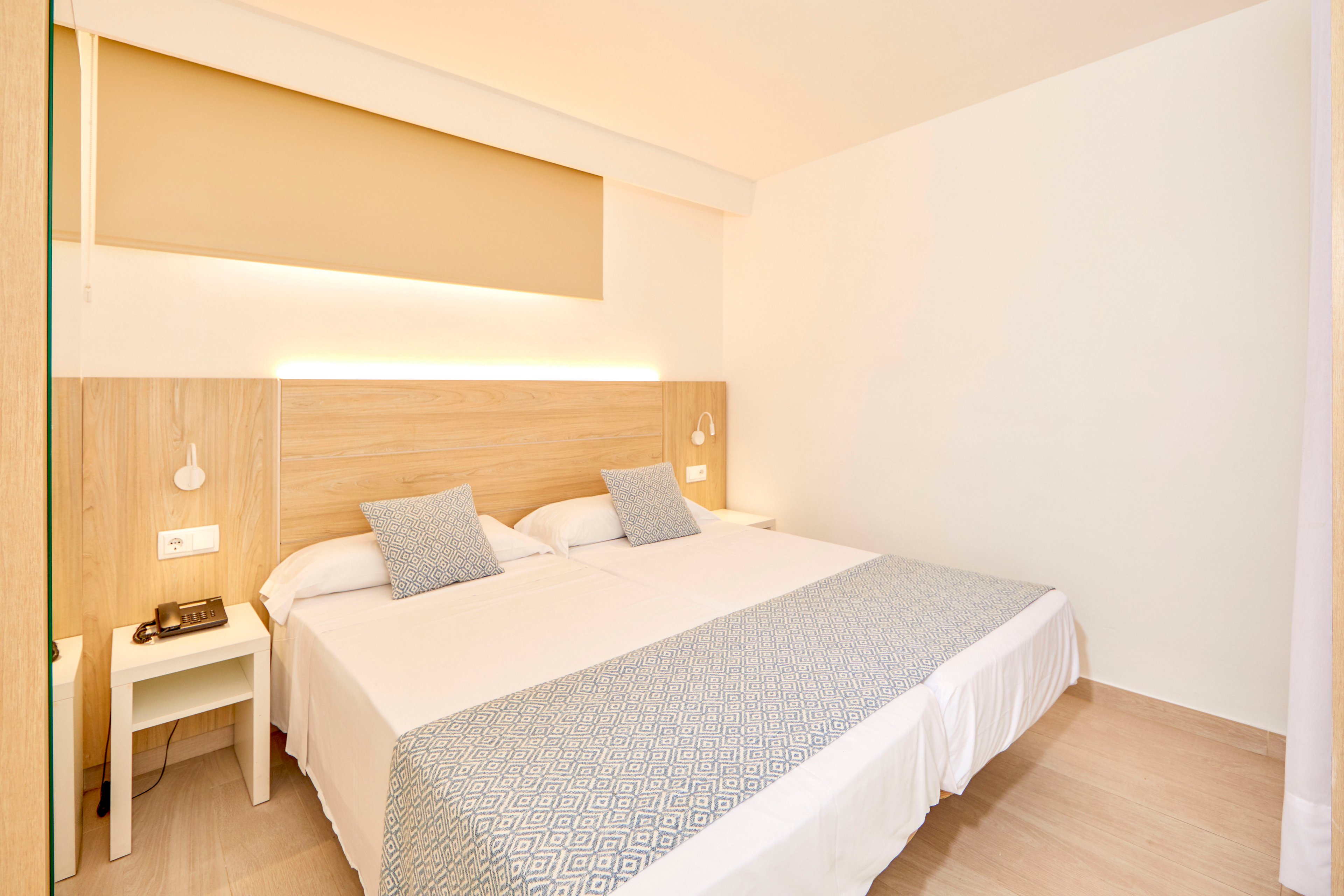 A bright, modern hotel bedroom features two twin beds pushed together with white linens and patterned gray throws. Light wood paneling behind the headboard includes integrated warm lighting and small hanging bedside lamps. A white nightstand holds a telephone and electrical outlets, emphasizing the room’s functional design. The overall aesthetic is minimalist, clean, and inviting with neutral tones and soft textures.