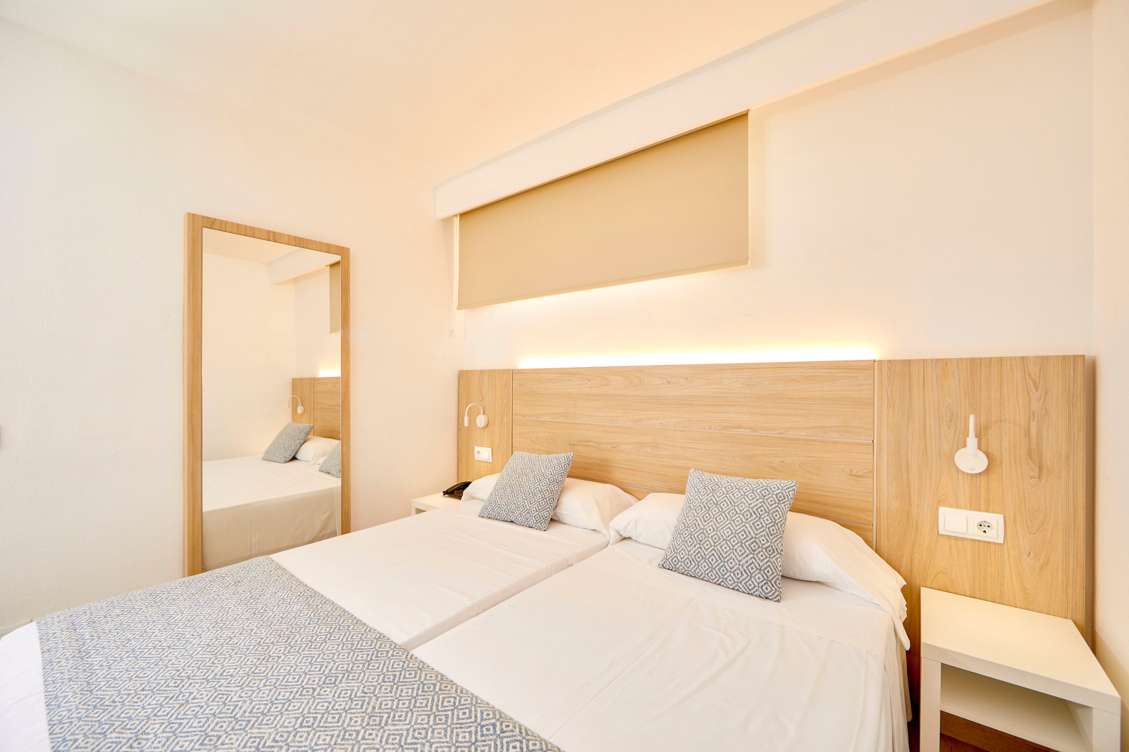 A bright, modern bedroom features two neatly made twin beds with white linens and light patterned throws. Soft wood tones on the headboard and bedside tables complement the neutral walls and warm ambient lighting. A large mirror reflects part of the room, enhancing the sense of space and cleanliness. The overall design is minimalist and inviting, suggesting a contemporary hotel or vacation rental interior.