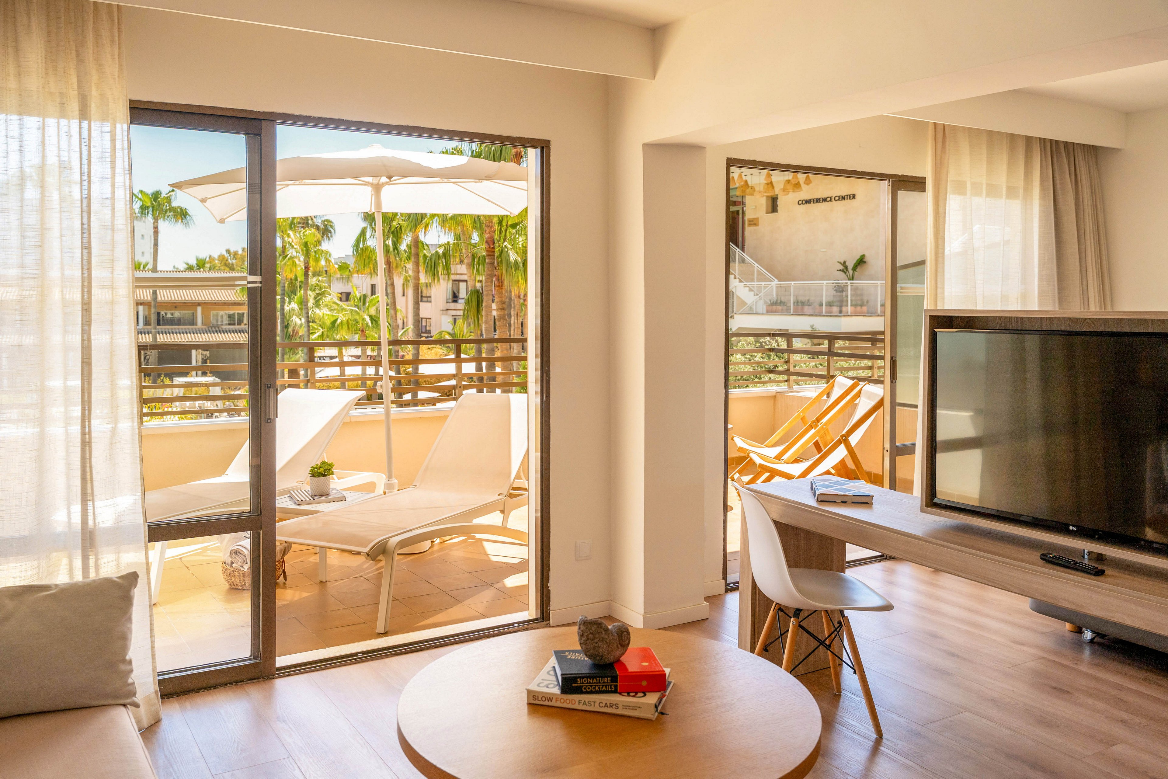 Bright sunlight fills a modern hotel suite with a large flat-screen TV and minimalist furnishings. Sliding glass doors open onto a spacious terrace with white sun loungers and umbrellas. Palm trees and neighboring resort buildings are visible outside, suggesting a warm vacation destination. Soft neutral tones and natural light create a relaxed, inviting atmosphere ideal for leisure and rest.