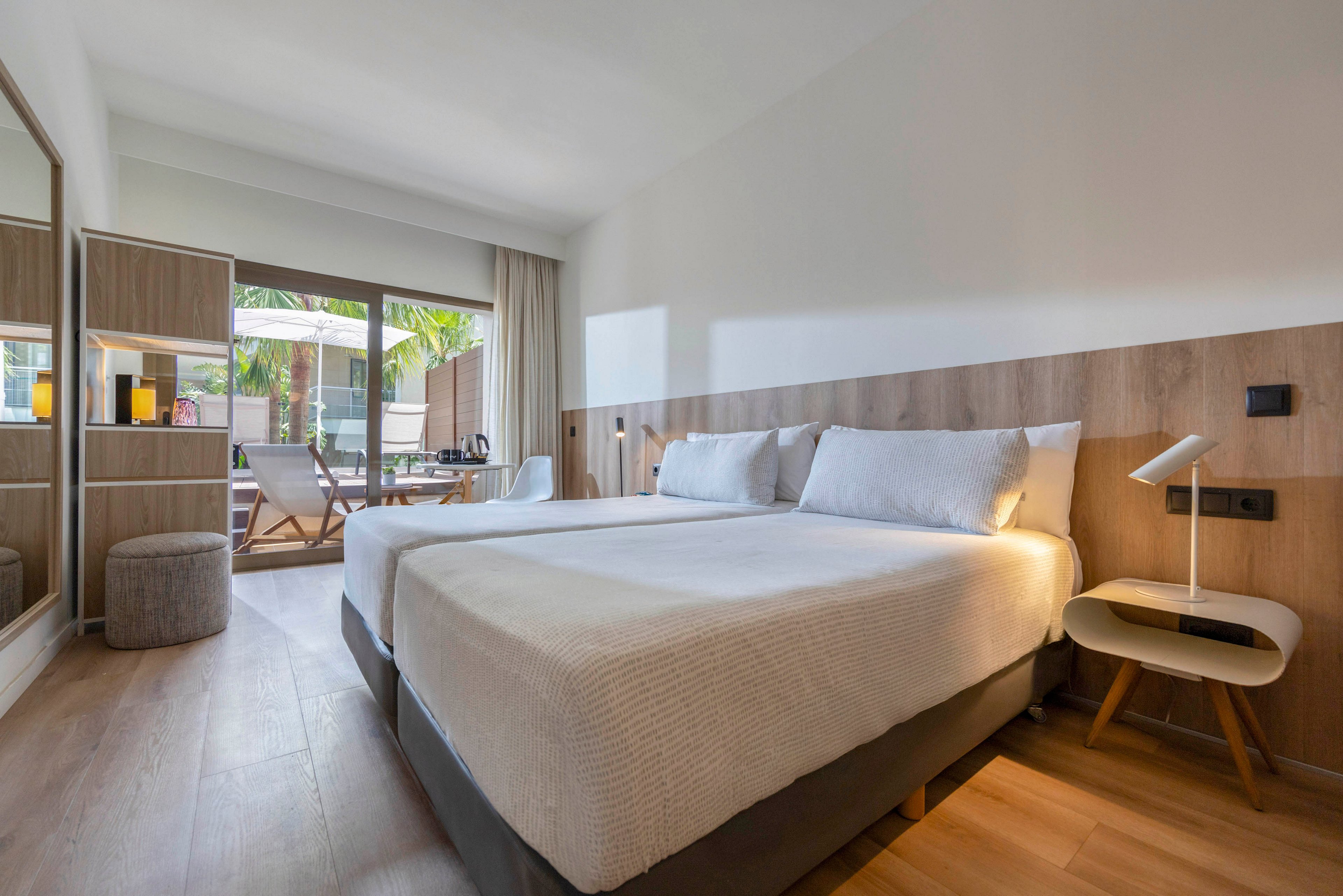 Bright contemporary hotel room featuring two neatly made beds with white linens. The space includes wooden furniture, a bedside lamp, and a cushioned stool, creating a clean and minimalist look. Large sliding glass doors open to a private terrace with outdoor seating and greenery. Natural light floods the room, highlighting the warm wood flooring and neutral color palette.