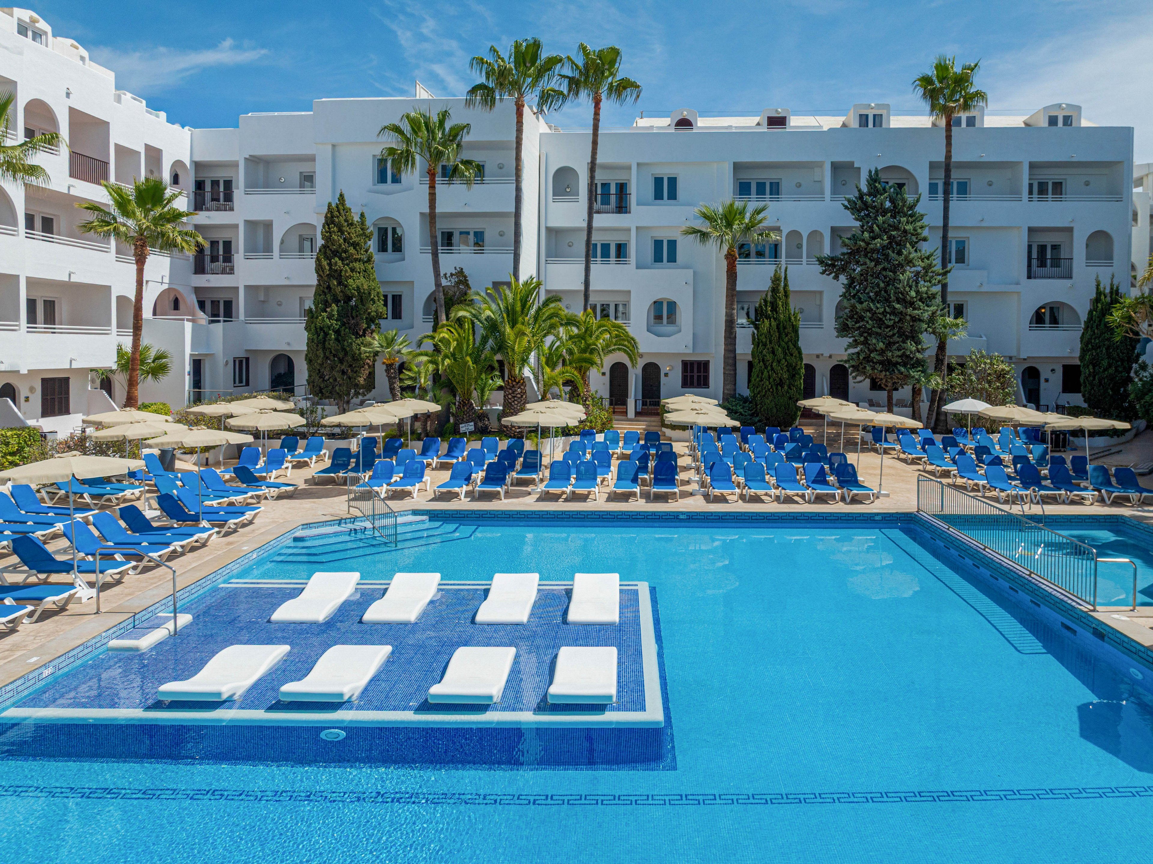 A large outdoor swimming pool is surrounded by rows of blue lounge chairs and beige umbrellas at a resort. White multi-story apartment-style buildings form the backdrop, with balconies overlooking the pool area. Palm trees and other greenery add a tropical feel to the bright, sunny scene. Several white floating loungers are arranged in the shallow section of the pool, emphasizing relaxation and vacation leisure.