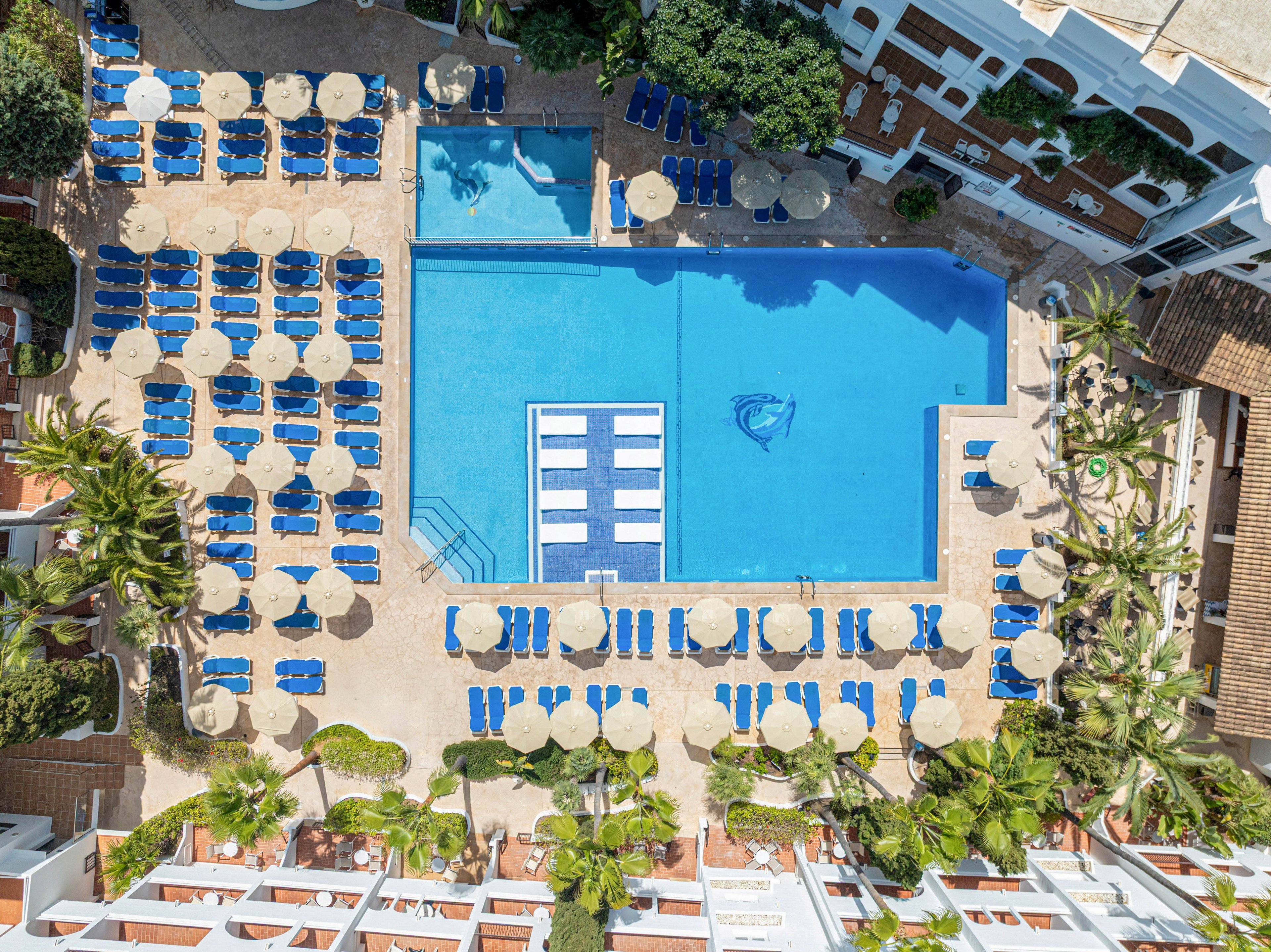 A large rectangular swimming pool is photographed from above at a sunny resort. Surrounding the pool are rows of blue lounge chairs and beige umbrellas arranged in a neat grid. Smaller pools and palm trees frame the main pool area, creating a tropical atmosphere. White buildings and pathways border the scene, emphasizing the organized, vacation-ready layout.