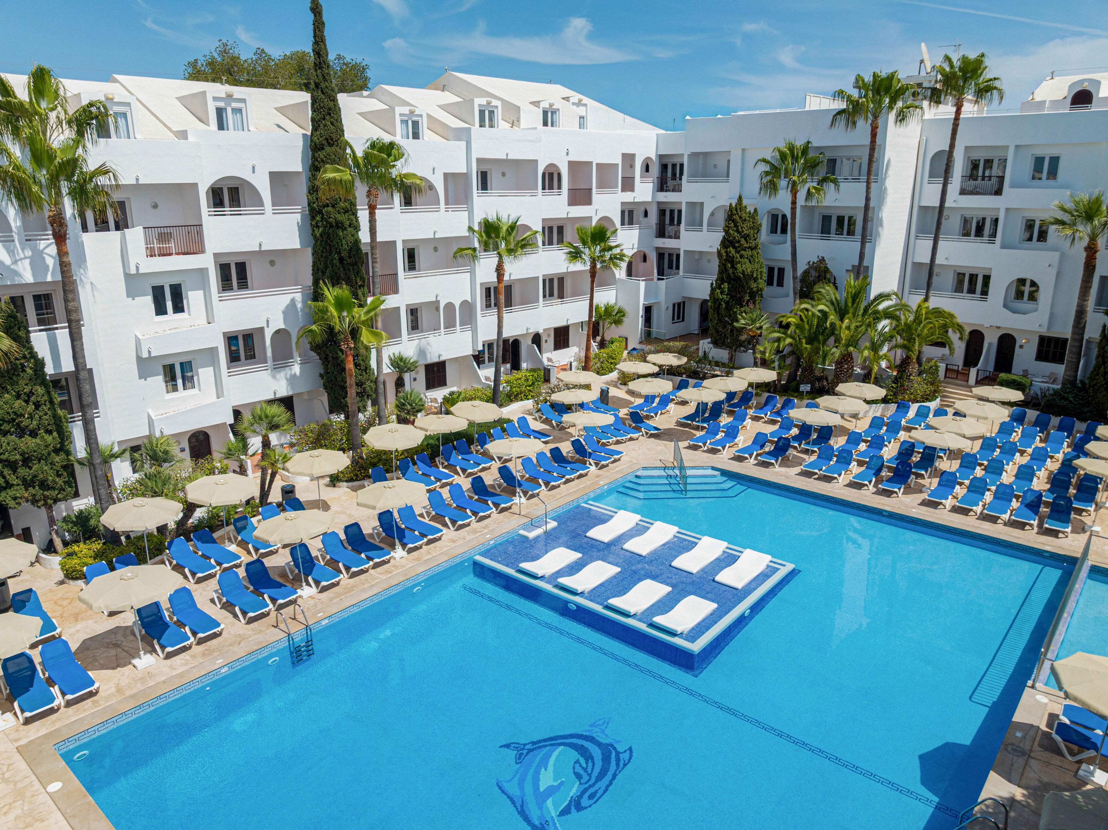 A large outdoor swimming pool is surrounded by rows of blue sun loungers and beige umbrellas. White multi-story resort buildings with balconies frame the pool area, with tall palm trees adding a tropical feel. The clear blue water features a decorative dolphin graphic on the pool floor and floating loungers in the center. The scene suggests a sunny vacation destination with ample seating for guests.
